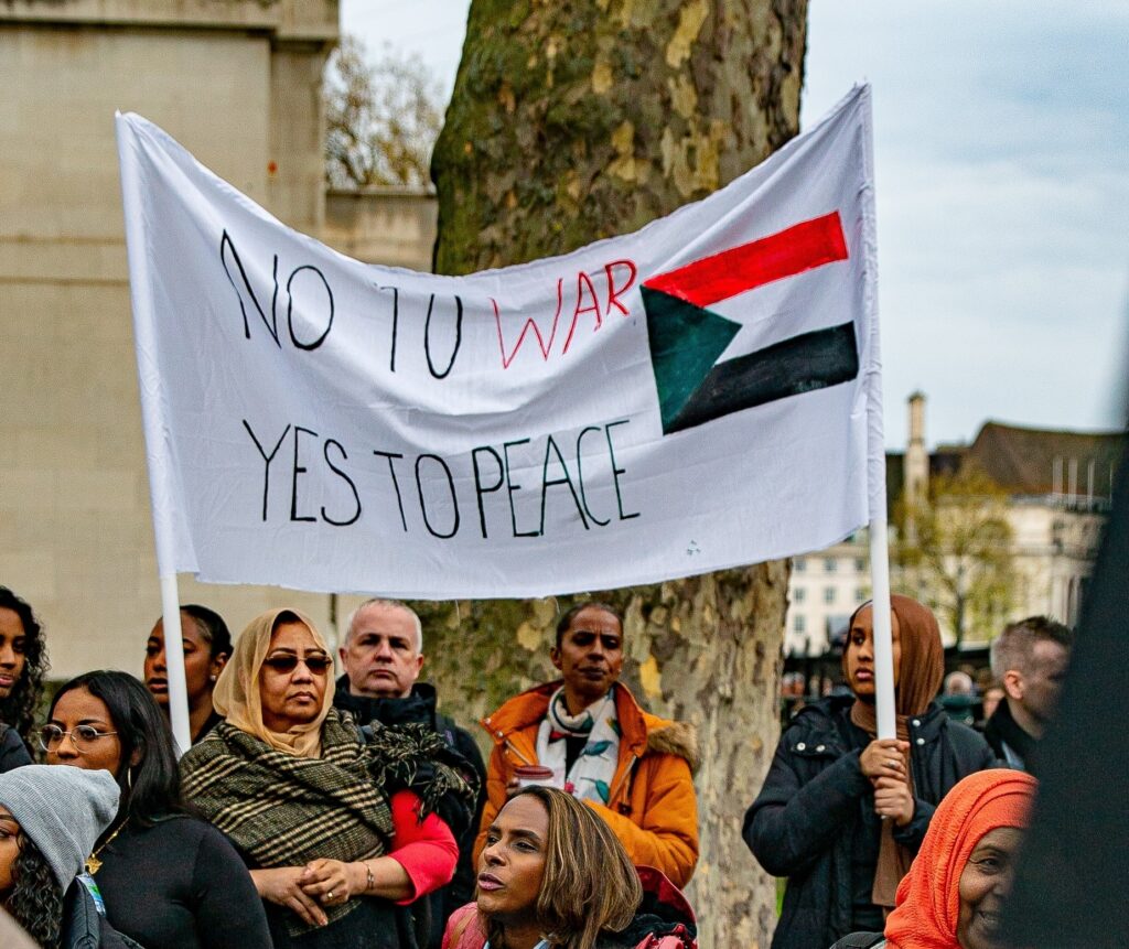 People holding a white sign that reads 'no to war, yes to peace' with the Sudanese flag on it