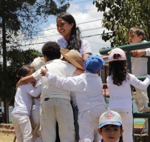 A group of children hugging their friend who is facing the camera laughing. The children are outside in blue sky.